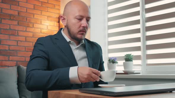 Portrait of young man in the suit drinks coffee and working with laptop on wooden work desk
