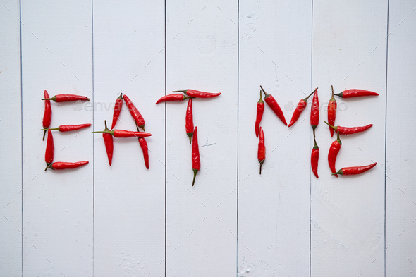 A word EAT ME formed with small red chilli peppers. Placed on white wooden table