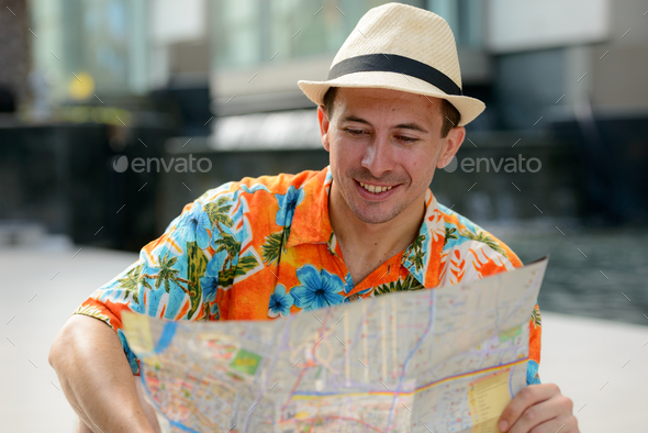 Happy young handsome tourist man reading map in the city outdoors