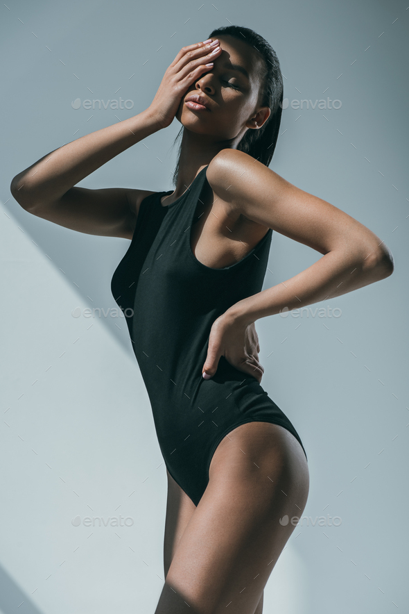 African American Trendy Woman With Slim Body Touching Her Face and Posing  in Black Bodysuit Stock Photo by LightFieldStudios