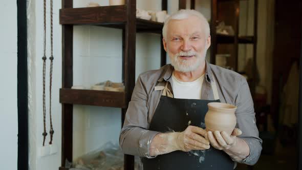 Portrait of Senior Potter in Apron Standing in Workshop Holding Pot Smiling alt
