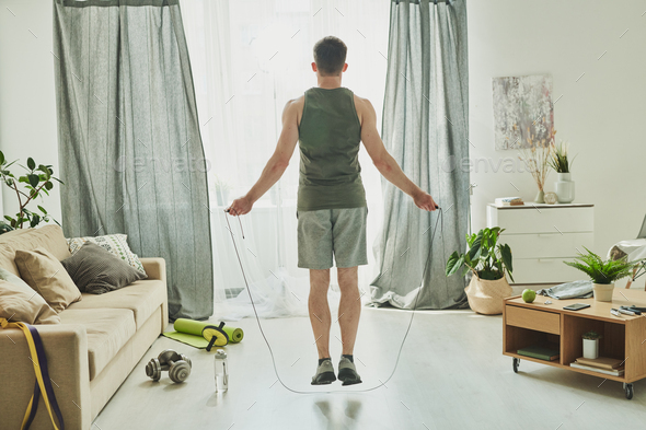 Back view of young man in activewear jumping with skipping-rope at home