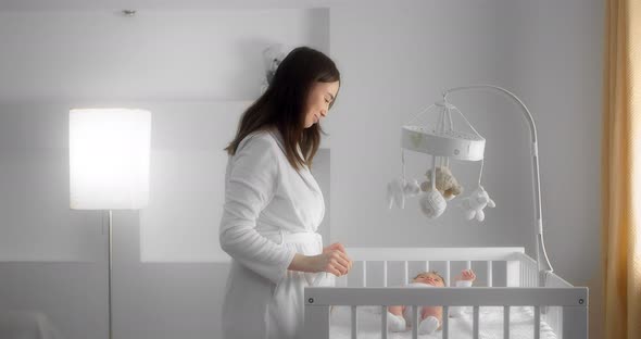 Mother in a White Coat Turns a Baby Mobile for a Newborn Baby Lying in a Crib
