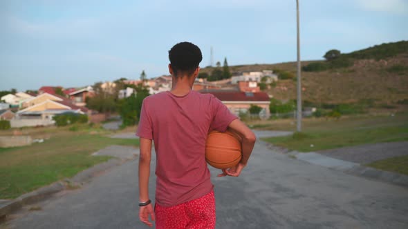 Rear View of Young Boy Walking with Basketball in Street
