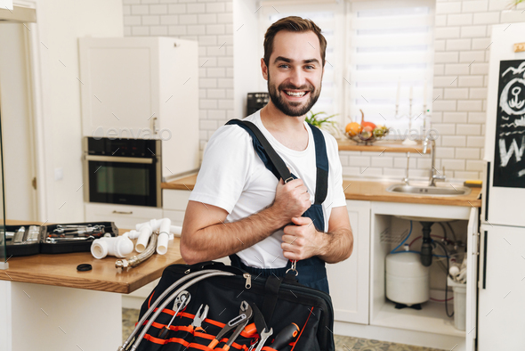 Image of plumber man smiling and holding bag with equipment in apartment