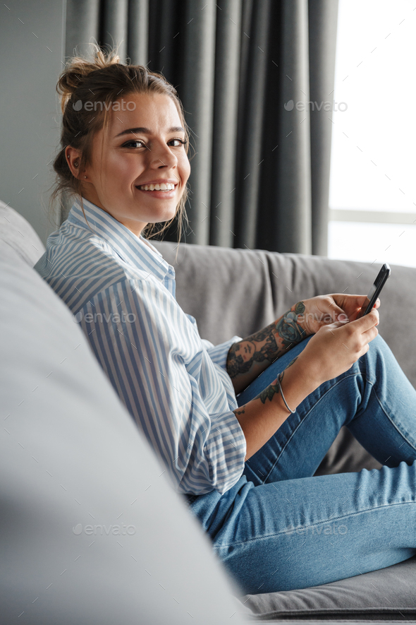Image of happy woman smiling and using cellphone while sitting on sofa