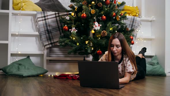 Beautiful girl lies with a laptop under a decorated Christmas tree on Christmas Eve