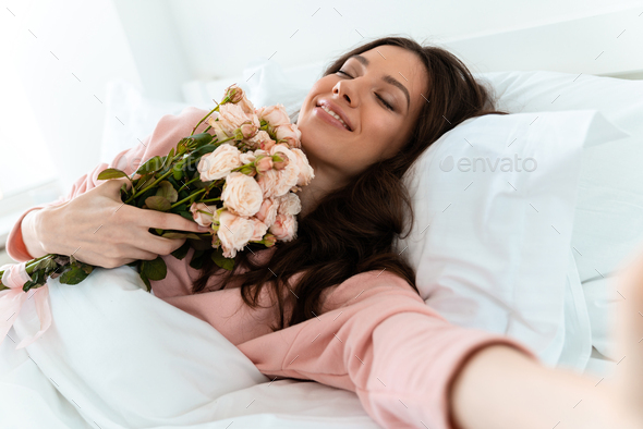 Pretty young woman in bed holding flowers. - Stock Photo - Images
