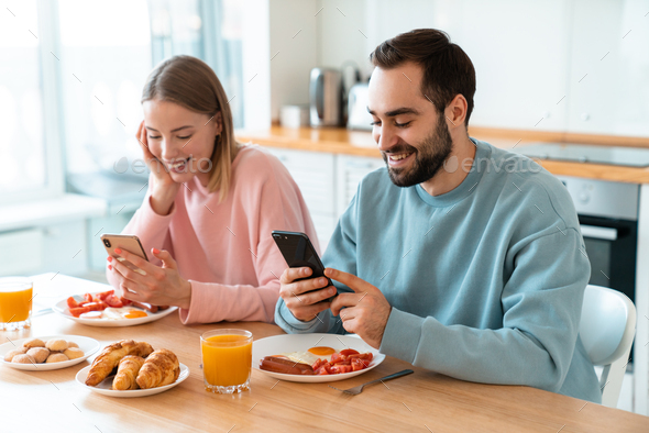 Portrait of young joyful couple using cellphones while having breakfast
