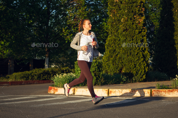 Young female runner, athlete is jogging in the city street in sunshine. Beautiful caucasian woman