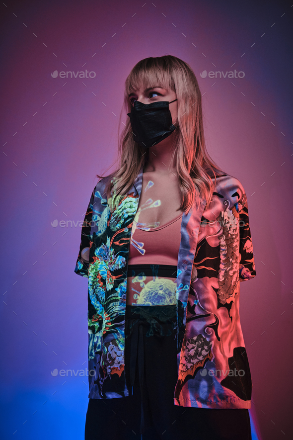 Young adult woman posing in a bright neon studio wearing protective medical mask - Stock Photo - Images
