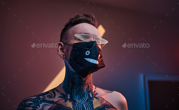 Inked male model posing in a neon studio with half-naked body, sunglasses and medical mask - Stock Photo - Images