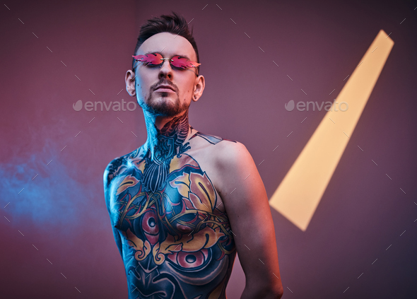 Inked male model posing in a neon studio with half-naked tattooed body and sunglasses. - Stock Photo - Images