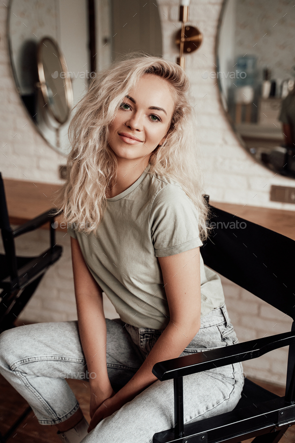 Confident blonde woman with curly hair sitting on a chair for make up in the studio full of mirrors - Stock Photo - Images