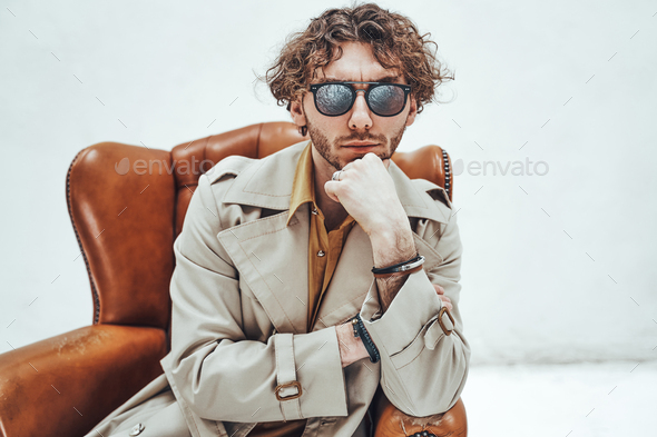 Handsome casual guy with curly hair posing in the bright studio while sitting on leather chair - Stock Photo - Images