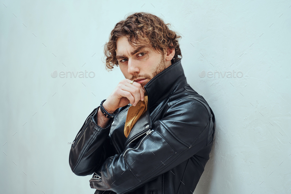 Daring and cool man with curly hair posing in leather coat in a bright studio - Stock Photo - Images