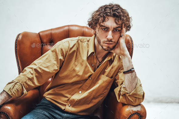 Handsome casual guy with curly hair posing in the bright studio while sitting on leather chair