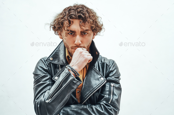 Young and and thoughtful male model with curly hair posing in a studio wearing leather jacket - Stock Photo - Images