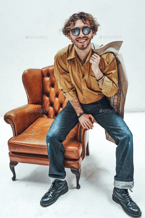 Handsome casual guy with curly hair posing in the bright studio while sitting on leather chair - Stock Photo - Images