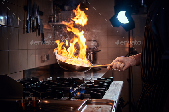 Cooker wearing gloves and apron frying flambe on a pan in a dark restaurant kitchen - Stock Photo - Images