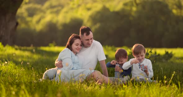 Loving Parents Hugging Sitting in the Field at Sunset and Looking Smiling at Two Sons Eating Ice alt
