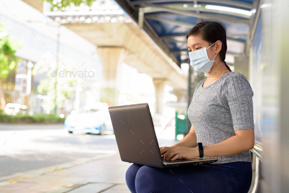 Young Asian woman with mask using laptop while sitting at the bus stop