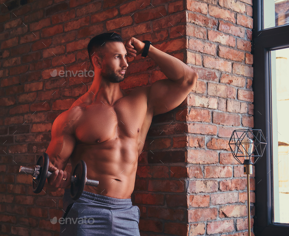 Tall stylish shirtless bodybuilder dressed in sports shorts in a room with  a loft interior. Stock Photo by fxquadro