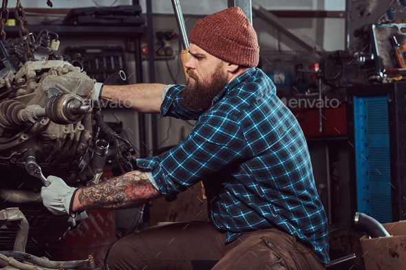 Brutal tattooed mechanic repairs the car engine which is raised on the hydraulic lift in the garage - Stock Photo - Images