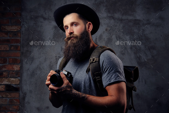 Handsome bearded traveler in a hat with a backpack and tattooed arms, holds a photo camera