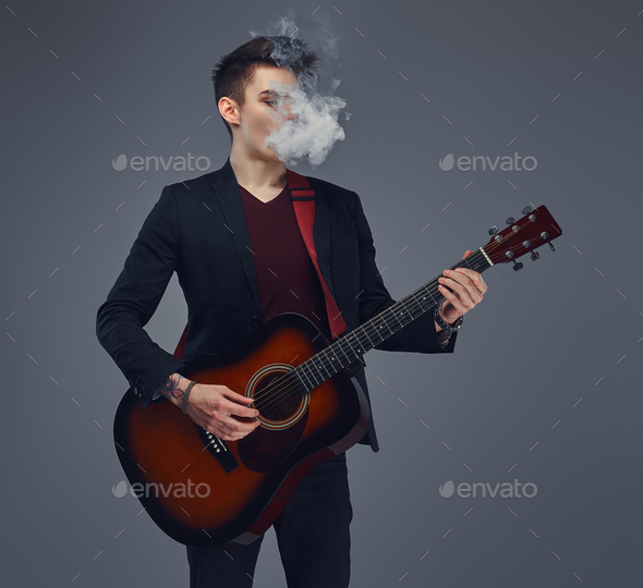 Young musician with stylish hair in elegant clothes exhales smoke while playing acoustic guitar.