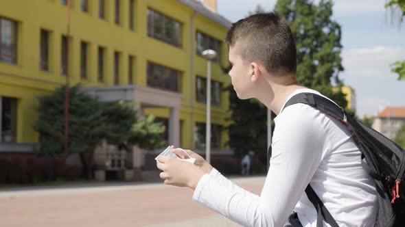 A Caucasian Teenage Boy Looks at a Face Mask in His Hands in Thought  a School in the Background