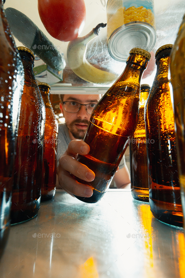 Caucasian man takes cold refreshing beer from out the fridge, inside view from fridge of hand
