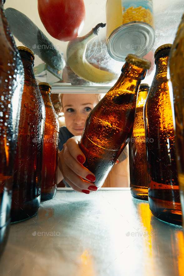 Caucasian woman takes cold refreshing beer from out the fridge, inside view from fridge of hand