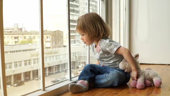 Little Girl Sits on Windowsill with Toy and Looks Out Window at Big City and Street at Sunset alt