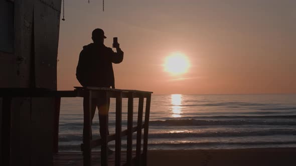 Silhouette of Young Man on Observation Deck Shooting Seaside Sunset on Phone