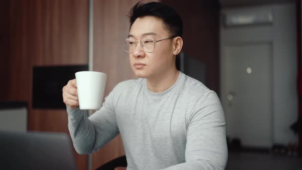 Asian serious man wearing eyeglasses working on laptop and drinking coffee alt