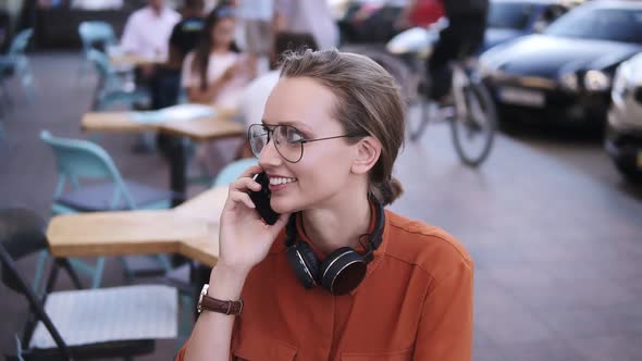 Close Up of Fair Hair Girl with Headphones on Her Neck Talking on Mobile Phone While Sitting in alt