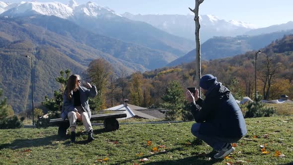 A Man Photographs a Young Woman Sitting on a Bench on a Hill in the Middle of the Beautiful Nature