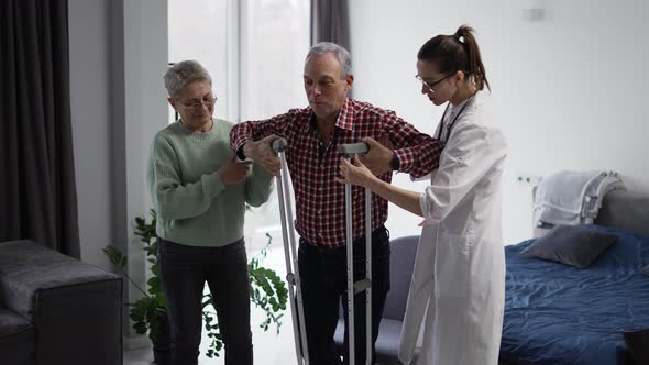 Old Man with Crutches Making First Steps with Support of Wife and Doctor alt