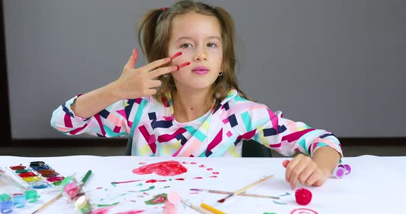 Cute Child Sitting at the Table and Drawing Stripe Camouflage on Her Face By Hand alt