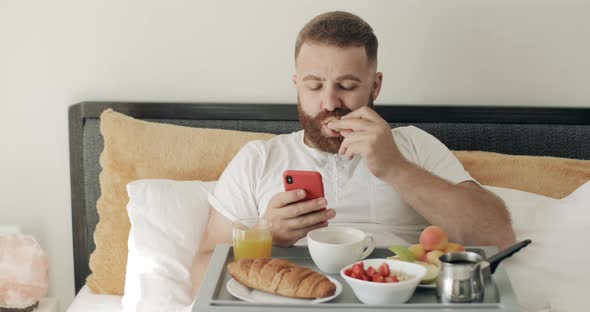 Serious Bearded Guy Using His Smartphone and Eating Apple While Sitting on Bed. Handsome Man in 30s alt