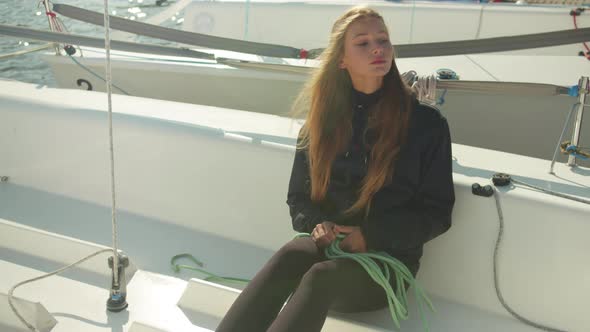 A Girl with Long Hair Sitting on the Deck of a White Sports Yacht Unwinds a Rope To Prepare the Boat alt
