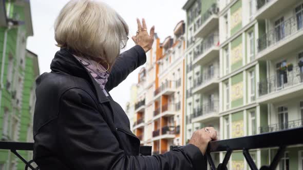 Side View Senior Woman Waving to Neighbors Standing on Urban City Balcony Outdoors alt