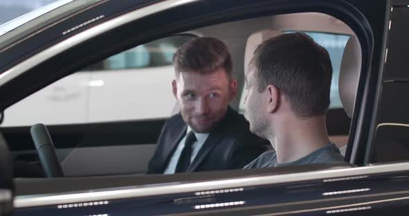 Side View of Confident Young Caucasian Man Taking Car Keys From Dealer and Starting Engine. Smiling alt