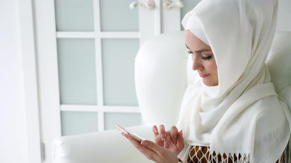 Attractive Muslim Woman in Hijab is Browsing Internet Pages in Smartphone Sitting on the Sofa alt