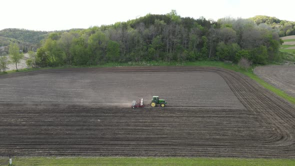 Drone view of farmer working to seed and fertilize field in rural valley. Hill covered in trees.