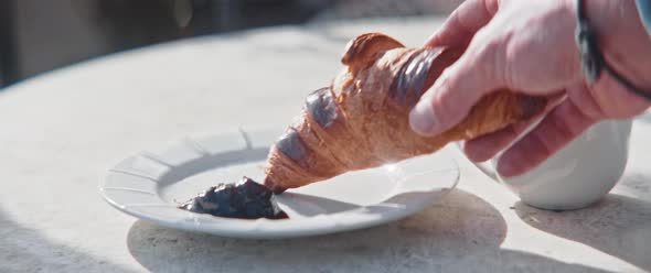 Hands of a man grabbing croissant and dip it in jam from a sunny balcony table closeup alt