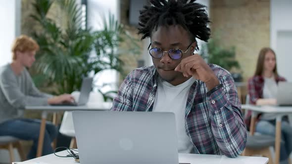 Focused Young African American Man Surfing Internet on Laptop with Blurred Colleagues Working Online alt