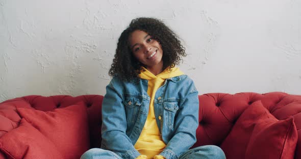 Cheerful African American Woman in Yellow Hoodie and Jeans Jacket Sitting on Red Sofa at Home