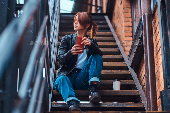 Redhead girl wearing trendy clothes sitting on stairs outside the cafe and holding a smartphone.